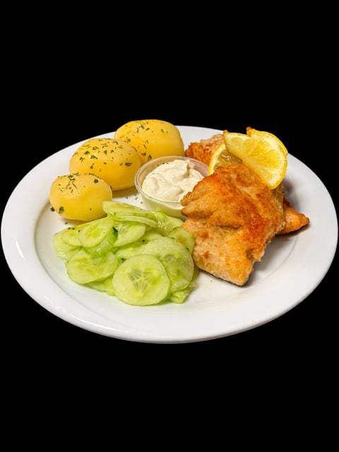 Plate of fried fish with boiled potatoes, cucumber slices, lemon wedges, and white dipping sauce on black background